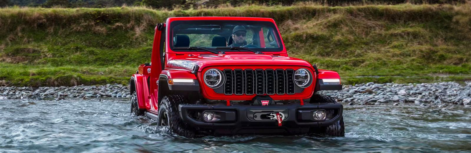 A front angle of a red 2024 Jeep Wrangler Rubicon crossing a large stream with water halfway up the wheels on the vehicle.