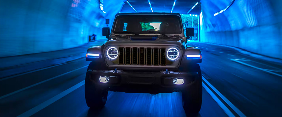 A front angle of a gray 2024 Jeep Wrangler Rubicon 4xe with its headlamps on, being driven through a…