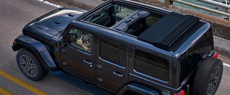 An angle from above of a black 2024 Jeep Wrangler High Altitude 4xe with its Sky One-Touch power sunroof fully open, being driven on a highway.
