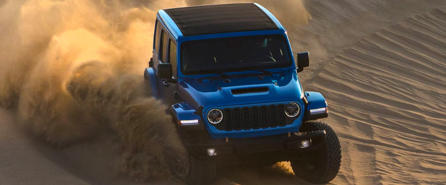 An angle from above of a blue 2024 Jeep Wrangler Rubicon 392 with black roof being driven down a sand dune.