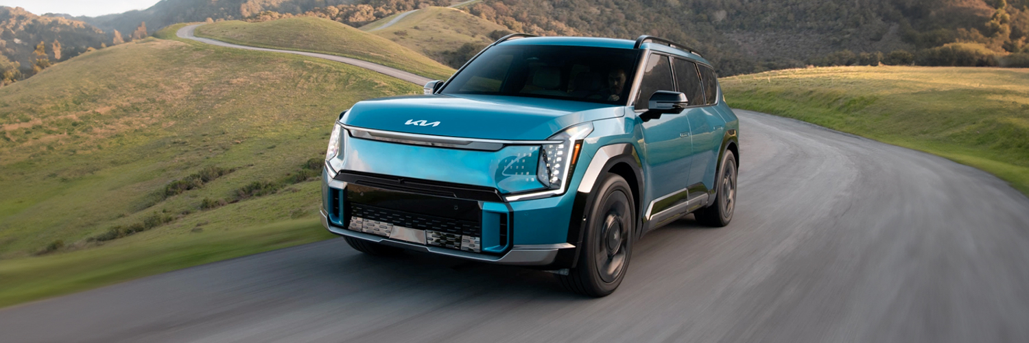 2024 Kia EV9 in blue, three-quarter front view, driving through a paved road in the hills