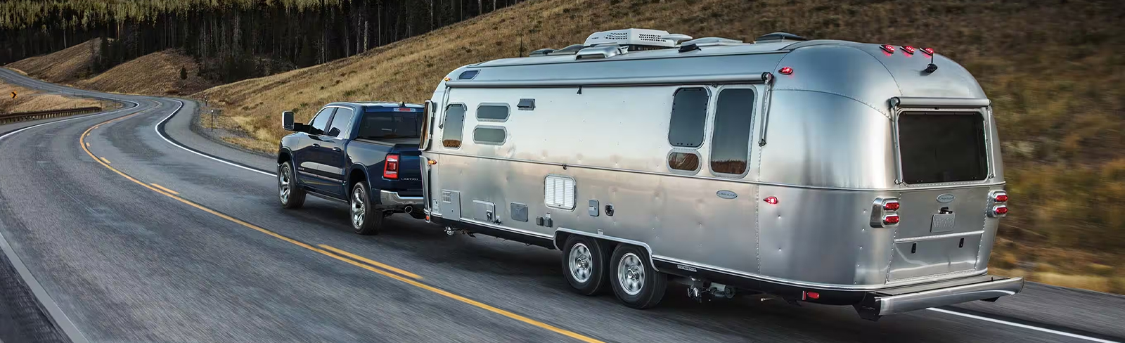 A black 2024 Ram 1500 Limited Crew Cab towing a large travel trailer as it is driven on a winding highway through the mountains.