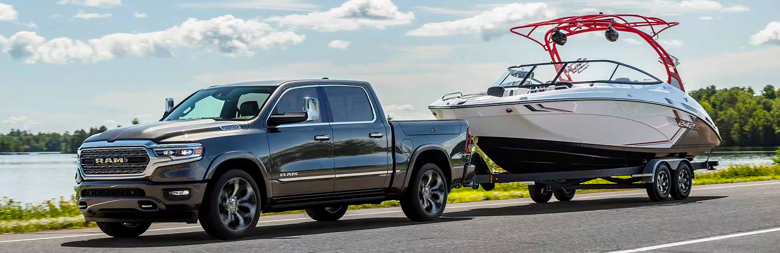 Display A silver 2024 Ram 1500 Limited Crew Cab towing a speedboat as it is driven down a highway beside a lake.