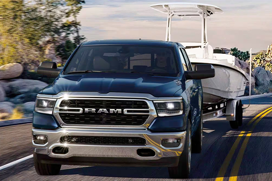 Display The front end of a blue 2024 Ram 1500 Big Horn Crew Cab towing a speedboat as it is driven down a winding highway in the mountains.