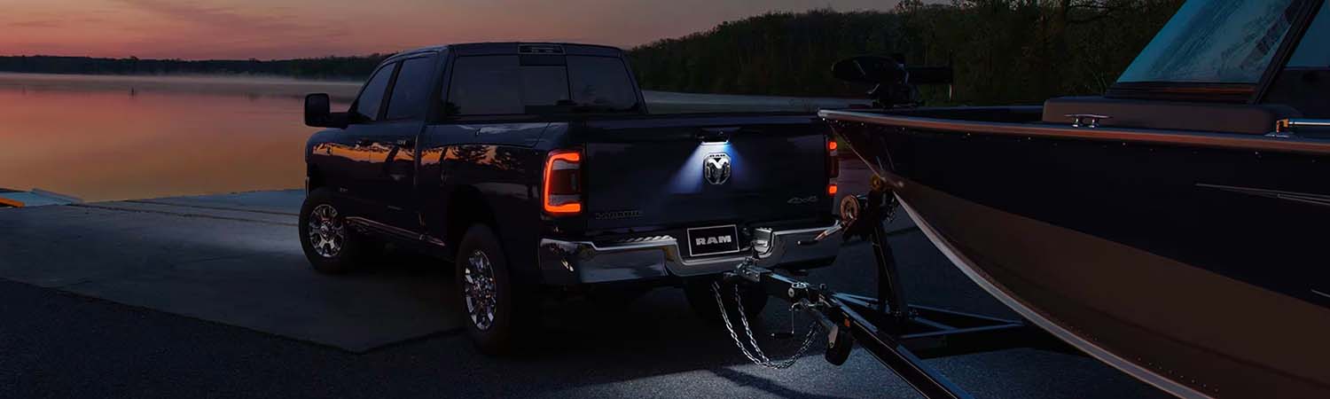 A rear angle of a 2024 Ram 2500 Laramie Crew Cab with its LED trailer hitch light and taillamps lit, towing a large speedboat on a boat ramp at night.