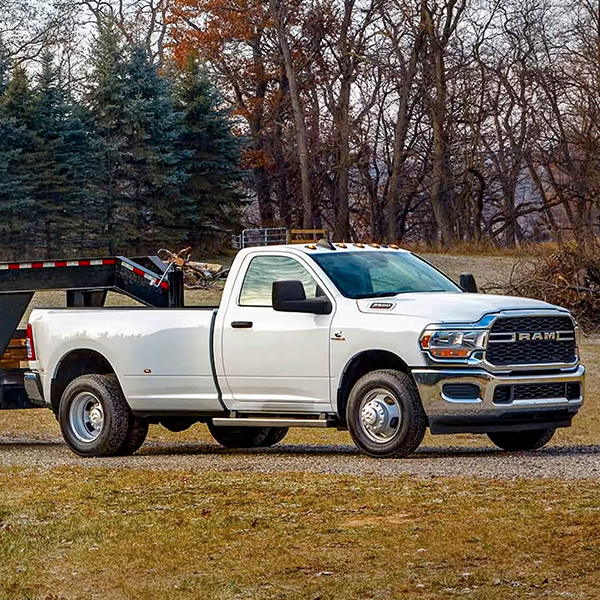 A white 2024 Ram 3500 Tradesman 4x4 Regular Cab towing a fifth wheel flatbed trailer loaded with an excavator and lumber.