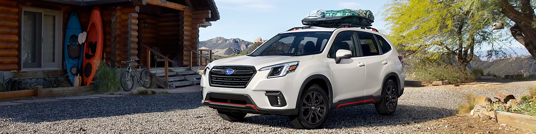 A white 2024 Subaru Forester with a cargo carrier on its roof parked in front of a log house
