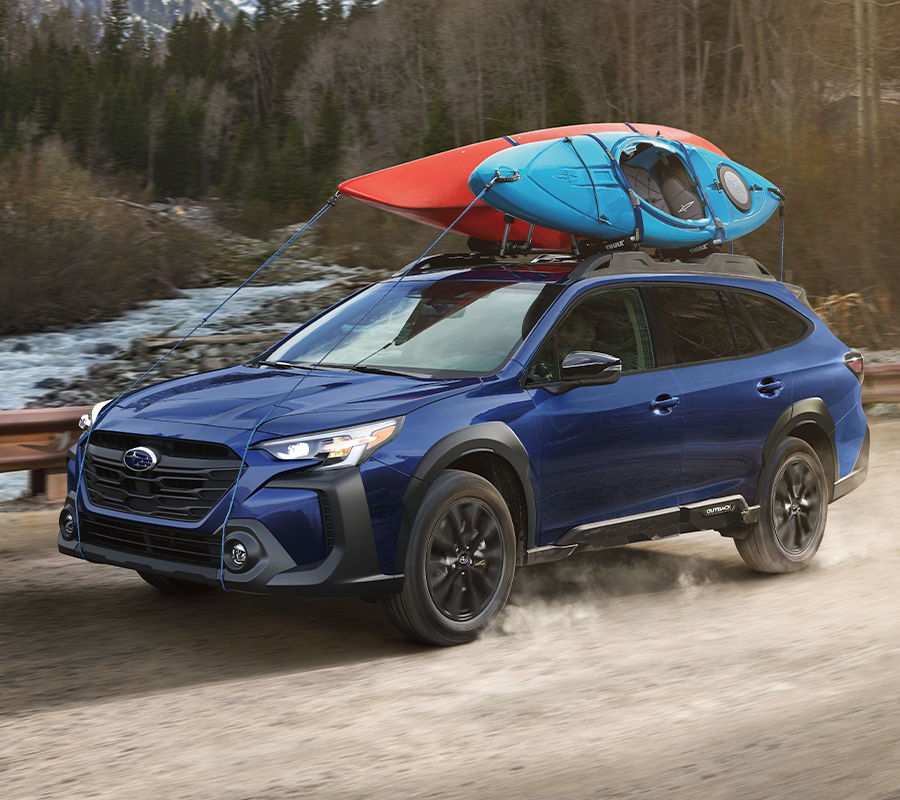 A Subaru Outback drives over a mountain river with 2 kayaks on its roof