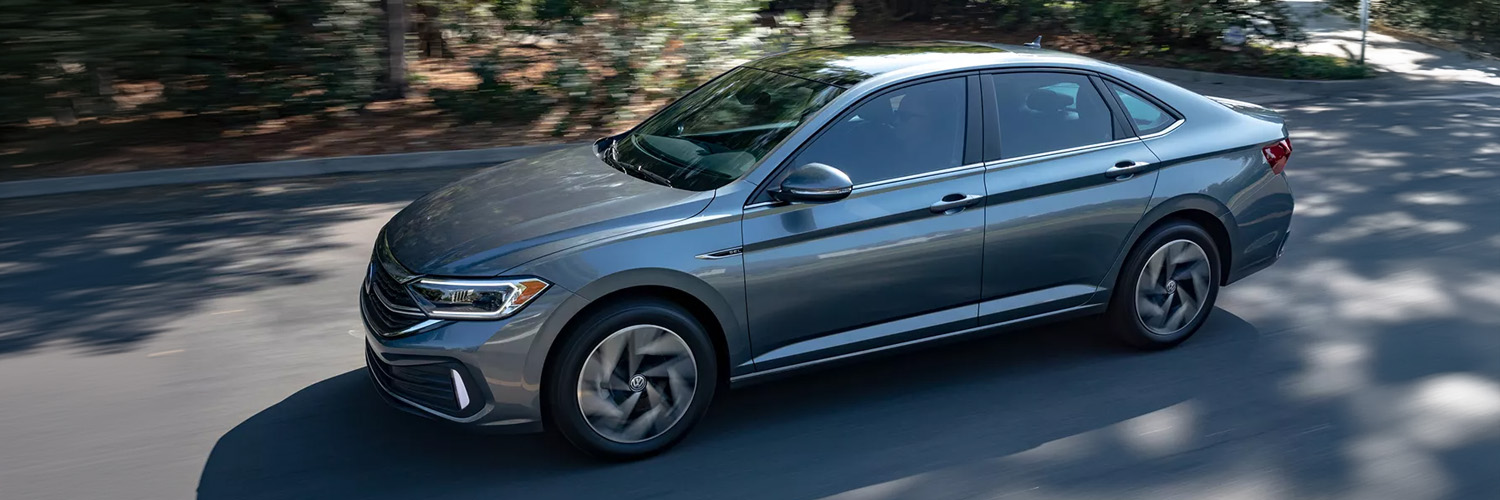 Side view of a Jetta in Platinum Gray Metallic driving on a tree lined road.