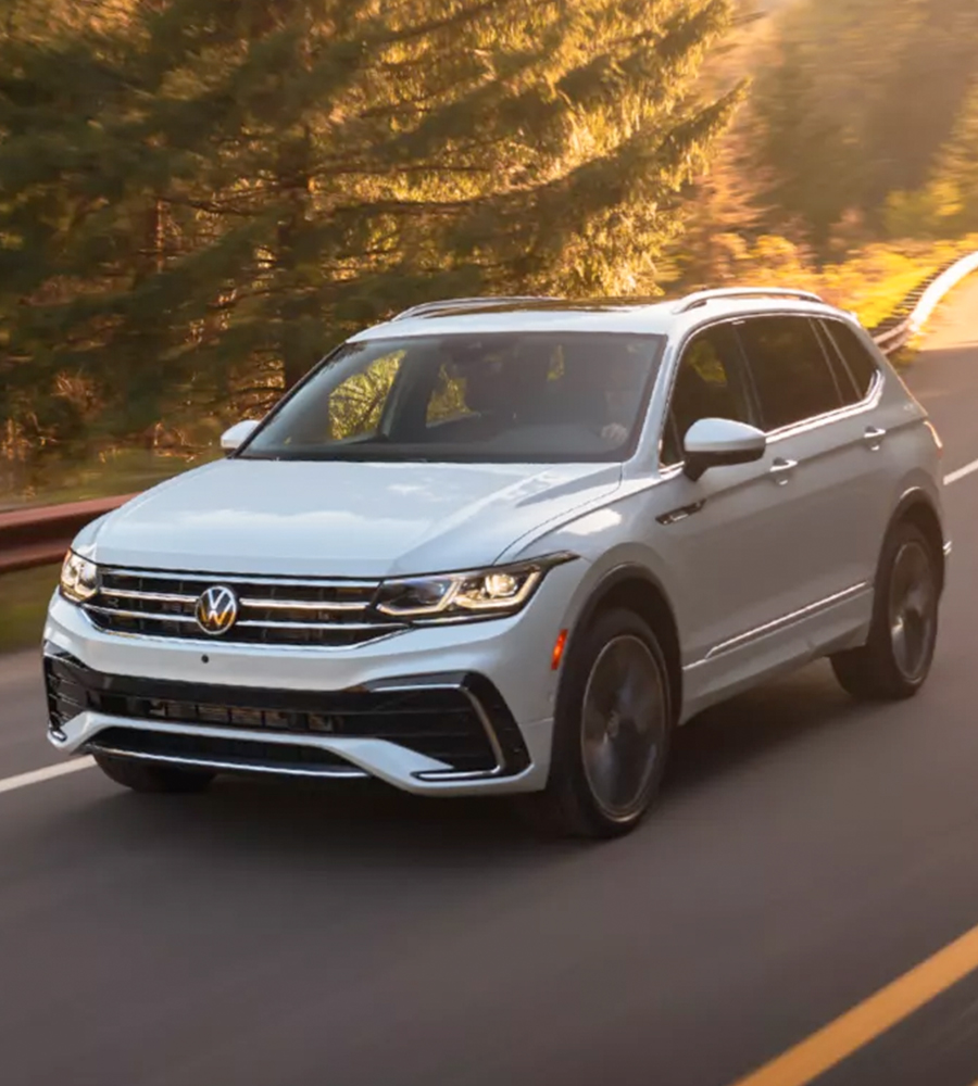 A Tiguan shown in Opal White drives down a winding, two-lane wooded road, as seen from the front driver side.