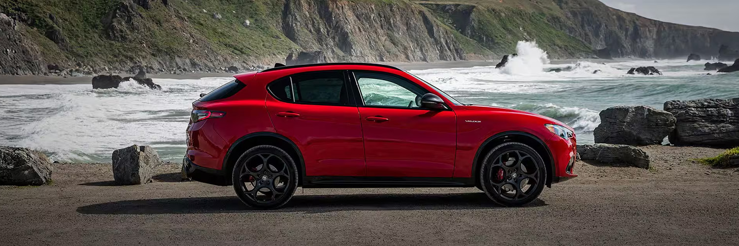 Side profile of a red 2025 Alfa Romeo Stelvio parked on a coastal road with ocean waves in the background.