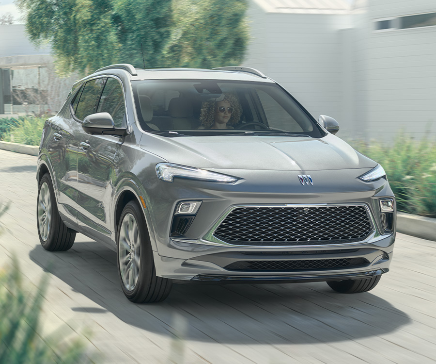 2025 Encore GX Avenir 7/8 Passengers side Front in Moonstone Gray Metallic. Vehicle on driveway with modern house and grasses.