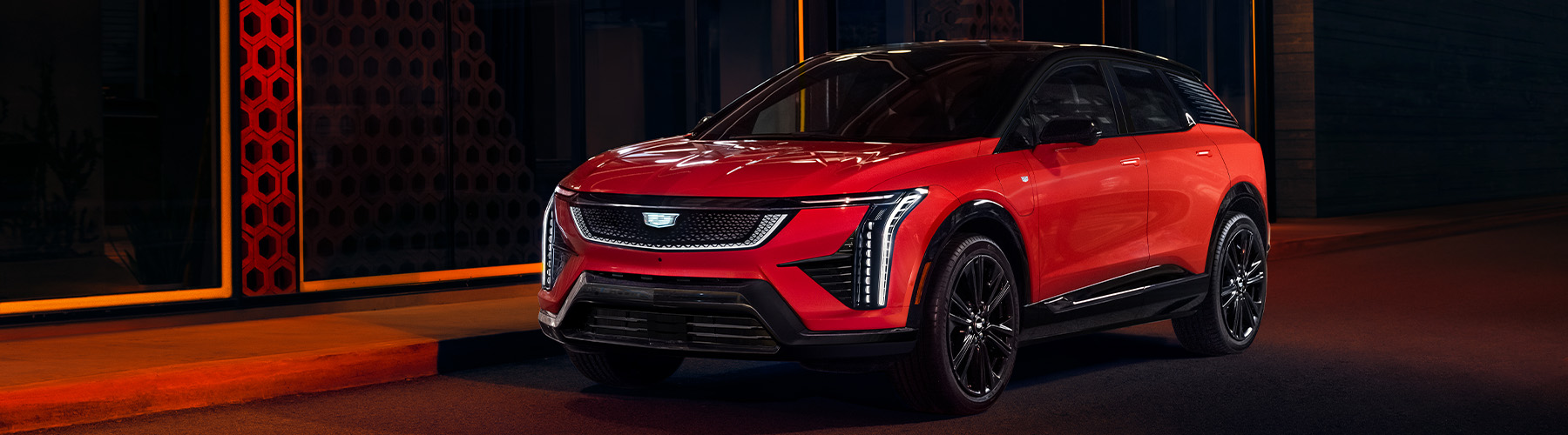 2025 Cadillac Optiq, Sport 2 (6MR26), exterior detail, red, Monarch Orange (GAG), 21 Inch wheel (RVU), parked, 3/4 driver front, night, dark, nighttime, red glow, outside building, modern, on street, city, headlamps on, lights.