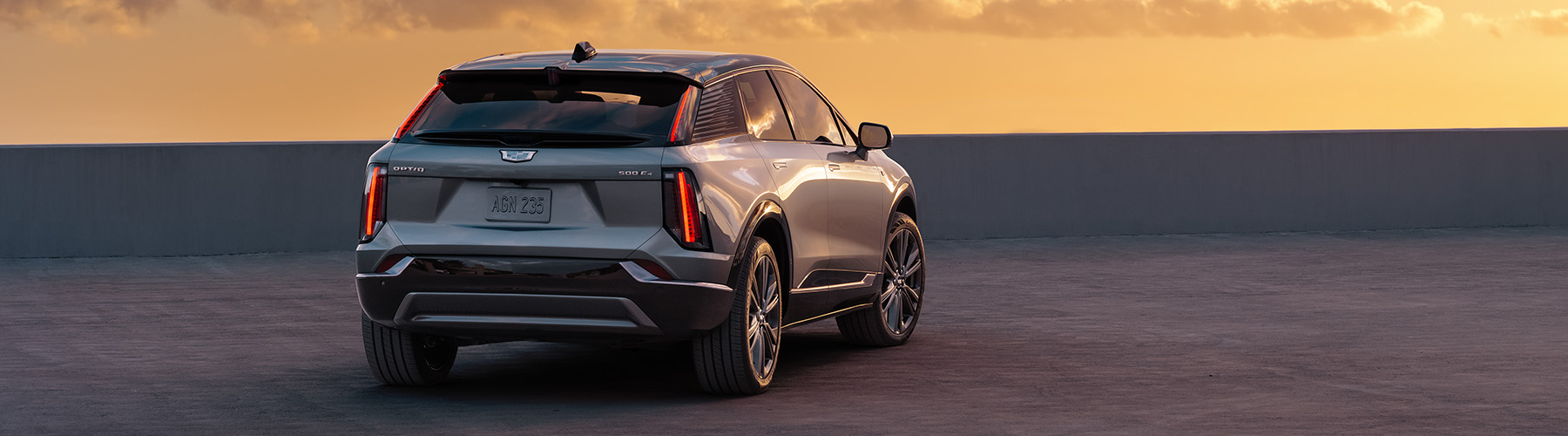 2025 Cadillac Optiq Sport 2 (6MR26) (1SH) in Argent Silver, 21-inch wheels RVU), Noir interior (EGW), 450 E4, 50/50 rear passenger side shot with sunset/sunrise in background, parked vehicle.