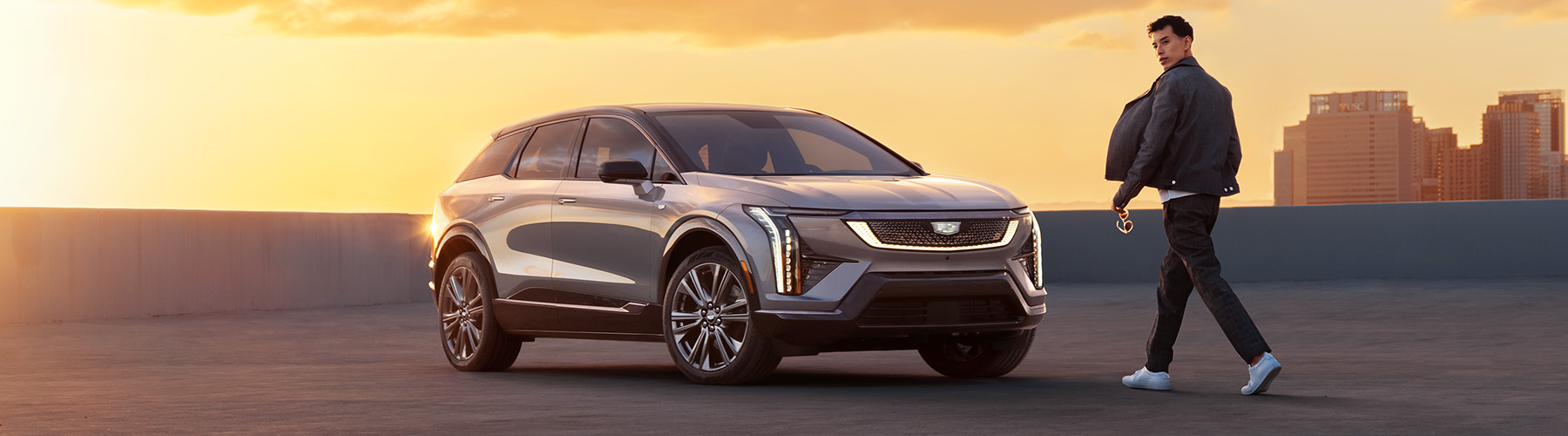 2025 Cadillac Optiq Sport 2 (6MR26) (1SH) in Argent Silver, 21-inch wheels RVU), Noir interior (EGW), 450 E4, 3/4 front driver side shot with sunset/sunrise in background, parked vehicle with city skyline, talent in frame.
