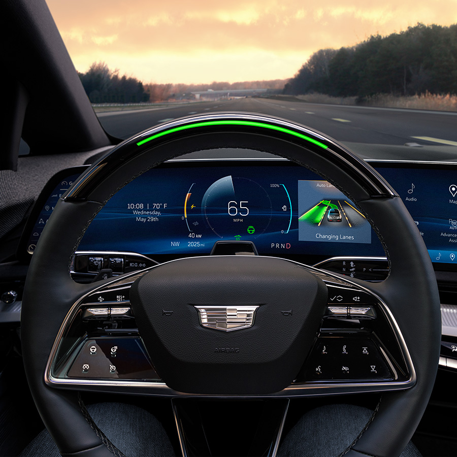 2025 Cadillac Optiq, Sport 2 (6MR26), interior detail, black upholstery, close up, NOIR (EGW), infotainment system, dashboard, through windshield, LED green steering wheel, controls, screen, sunset, clouds, in motion, on road, paved, trees, forest.