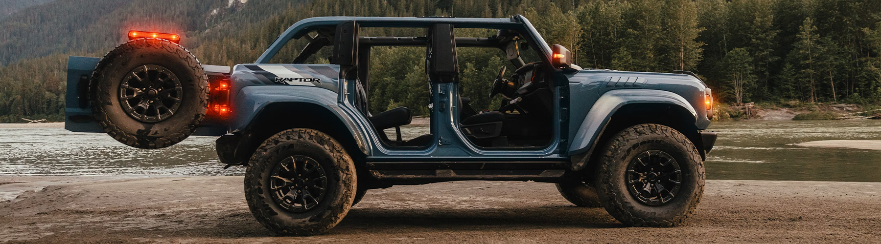 A 2025 Ford Bronco® SUV with the doors removed and the tailgate open, parked on a mud flat near a mountain lake