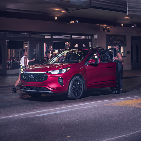 Three people near a 2025 Ford Escape® SUV in Rapid Red Metallic Tinted Clearcoat (extra-cost paint) parked near a theatre