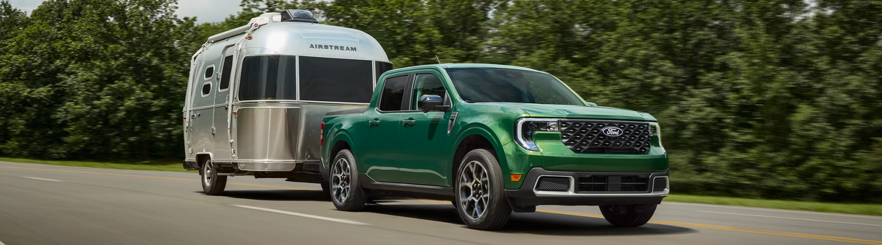 A 2025 Ford Maverick® pickup pulling an Airstream travel trailer along a highway, with a forest behind