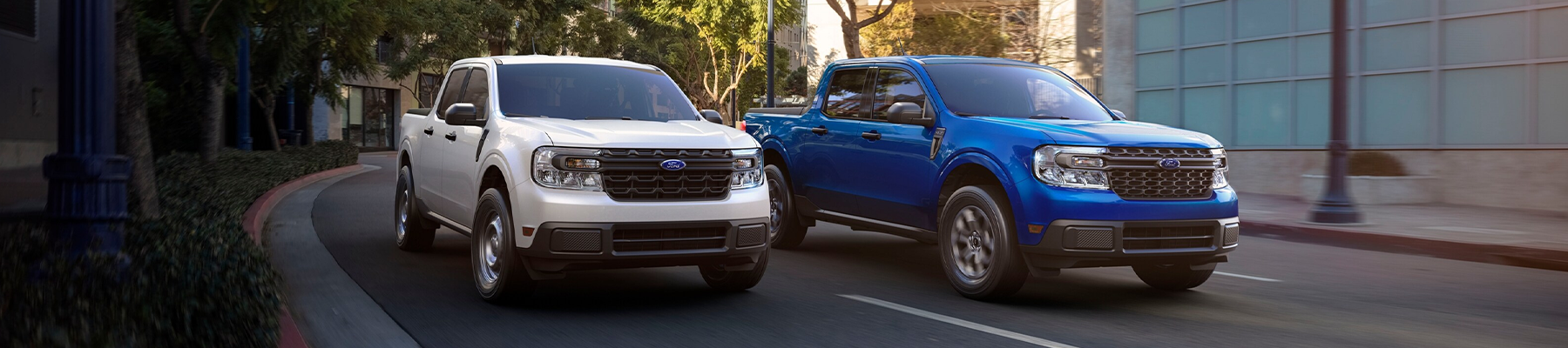 Two 2024 Ford Maverick® trucks in Oxford White and Atlas Blue Metallic are being driven on a tree-lined city road