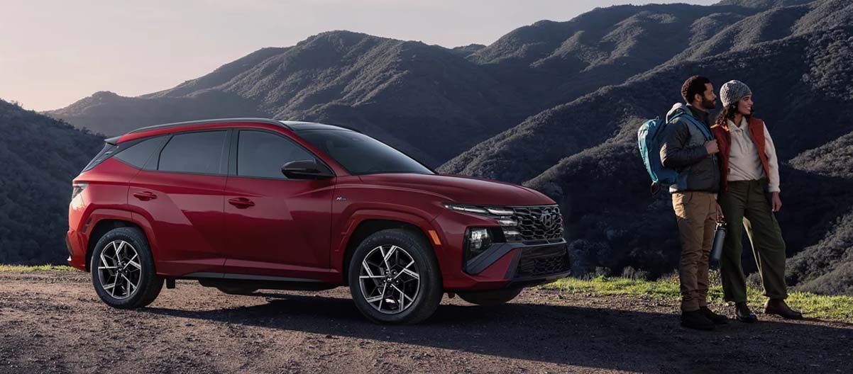 Red 2025 Hyundai Tucson Hybrid parked along a mountainous road with a happy couple nearby