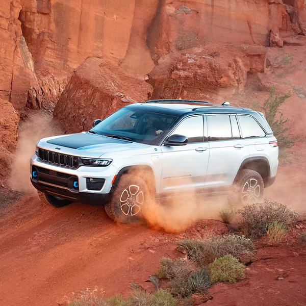 Display A white 2025 Jeep Grand Cherokee Trailhawk 4xe with black hood insert, kicking up sand as it's driven off-road beside huge red boulders.