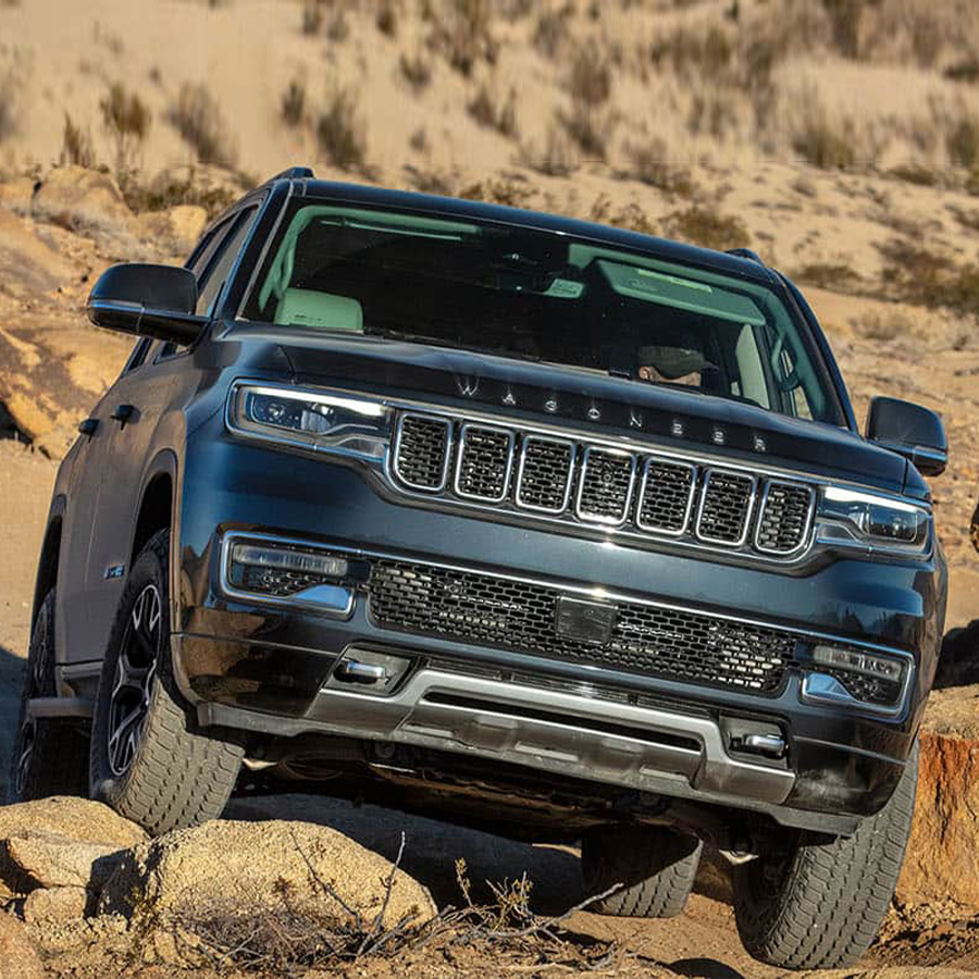 Display A front end angle of a gray 2025 Jeep Wagoneer Series III ascending a rocky hill with the tires crawling over an uneven surface of different size boulders.