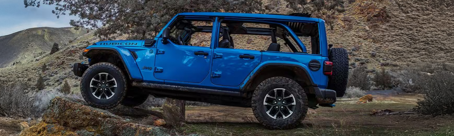 Display A driver-side profile of a blue 2025 Jeep Wrangler 4xe Rubicon X 4xe parked off-road, with its front tires elevated on a raised rock formation.