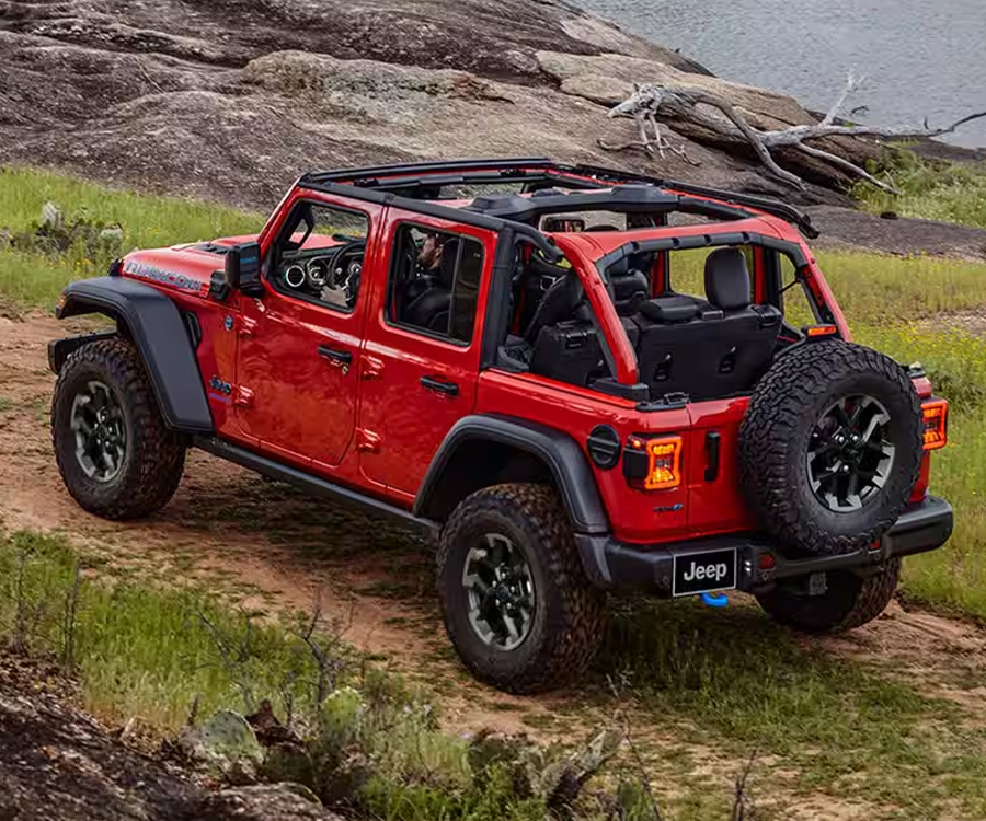 A red 2025 Jeep Wrangler Rubicon 4xe with its roof removed, traveling on a trail off-road, beside a lake.