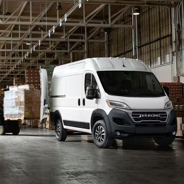A passenger-side front angle of a white 2025 Ram ProMaster 2500 SLT Cargo Van High Roof, parked in a warehouse with its rear doors fully open and a forklift with a pile of boxes being driven toward the back of the van.