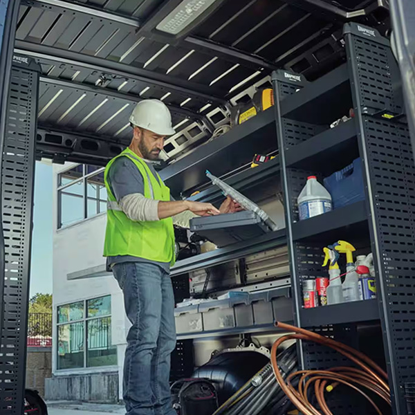 Display The 2025 Ram ProMaster Cargo Van High Roof with its passenger-side sliding door and rear doors open, displaying shelves full of tools and equipment and a technician working on a laptop.