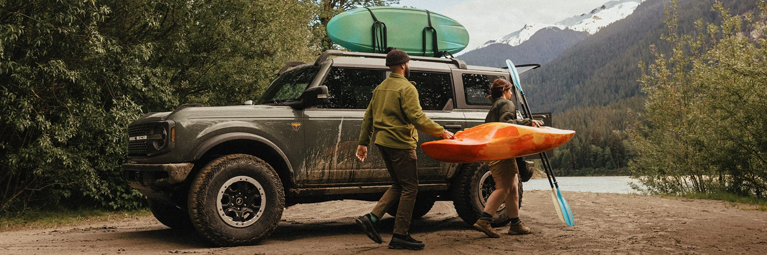 A man and woman carrying a kayak and paddle away from a 2026 Ford Bronco® SUV parked on a mud flat