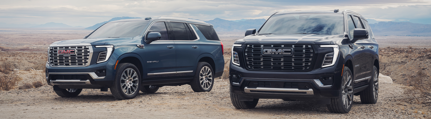 2026 GMC Yukon Denali Blue and Denali Ultimate in Black front driver side profile on a dirt road with clouds overhead