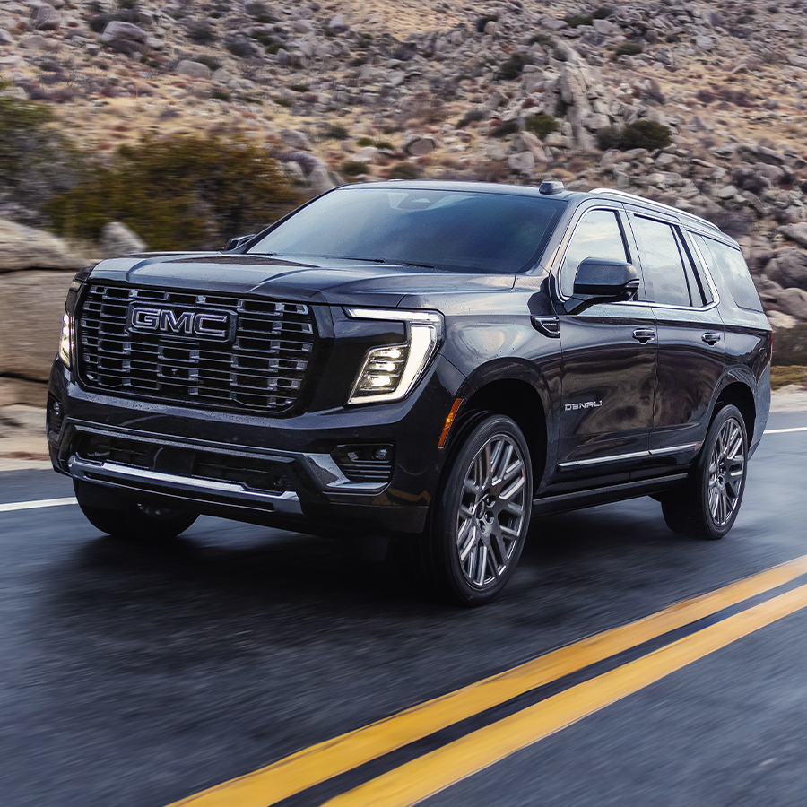 2026 GMC Yukon Denali Ultimate in Rush Metallic 3/4 Driver side view profile driving down paved road with mountains in the background
