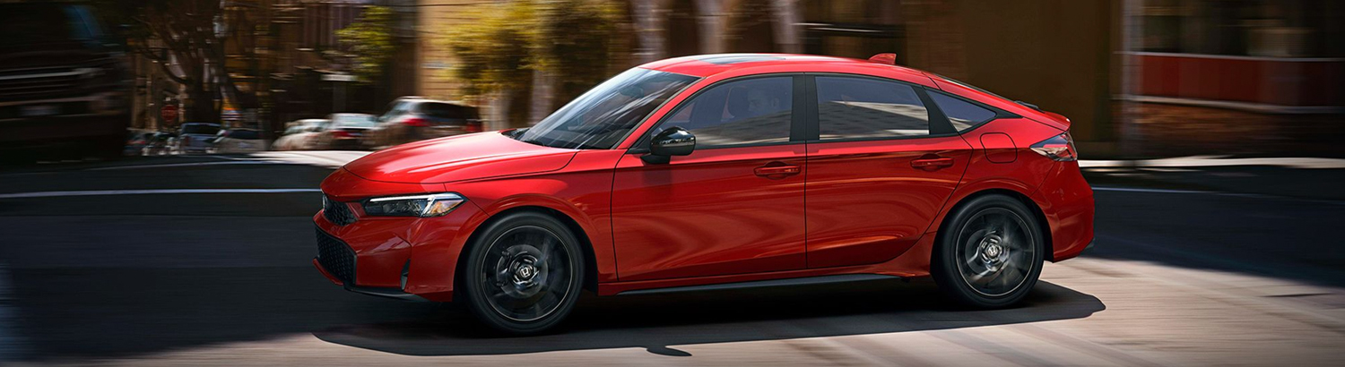 2026 Honda Civic Hatchback Sport Hybrid in Rallye Red driving through intersection of urban environment