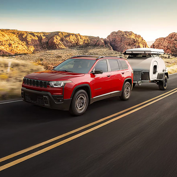 Red 2026 Jeep Cherokee towing a small camper trailer through a desert highway.