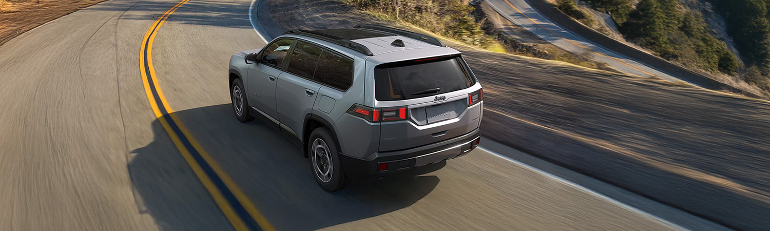 Rear view of a silver 2026 Jeep Cherokee driving along a winding mountain road.