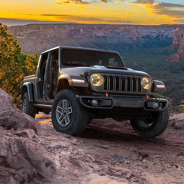 Display A gray 2026 Jeep Gladiator Mojave ascending a rocky trail off-road, high in the mountains at sunset.