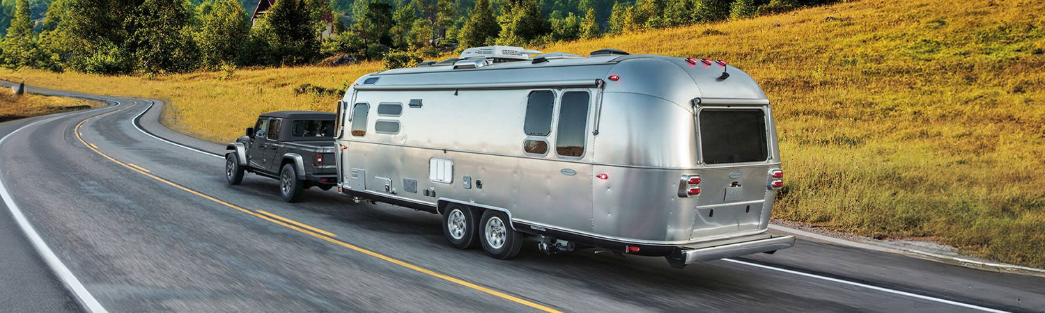 Display A gray 2026 Jeep Gladiator Sport S traveling down a highway, towing a large travel trailer, with mountains in the background.
