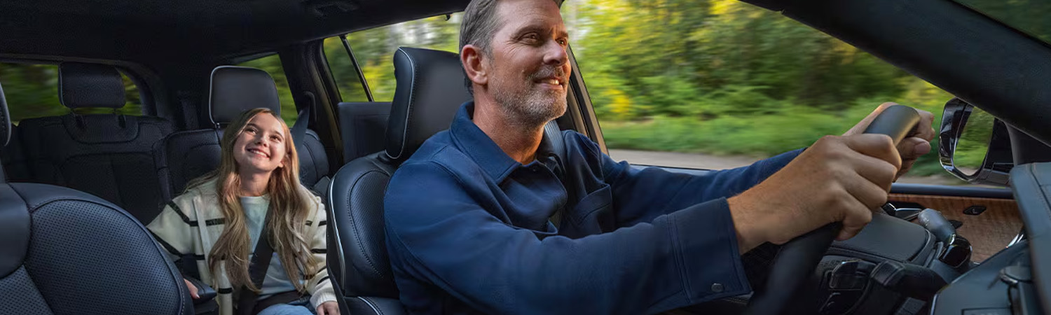 Interior shot of a 2026 Jeep Grand Cherokee with a dad driving and his daughter smiling in the back seat