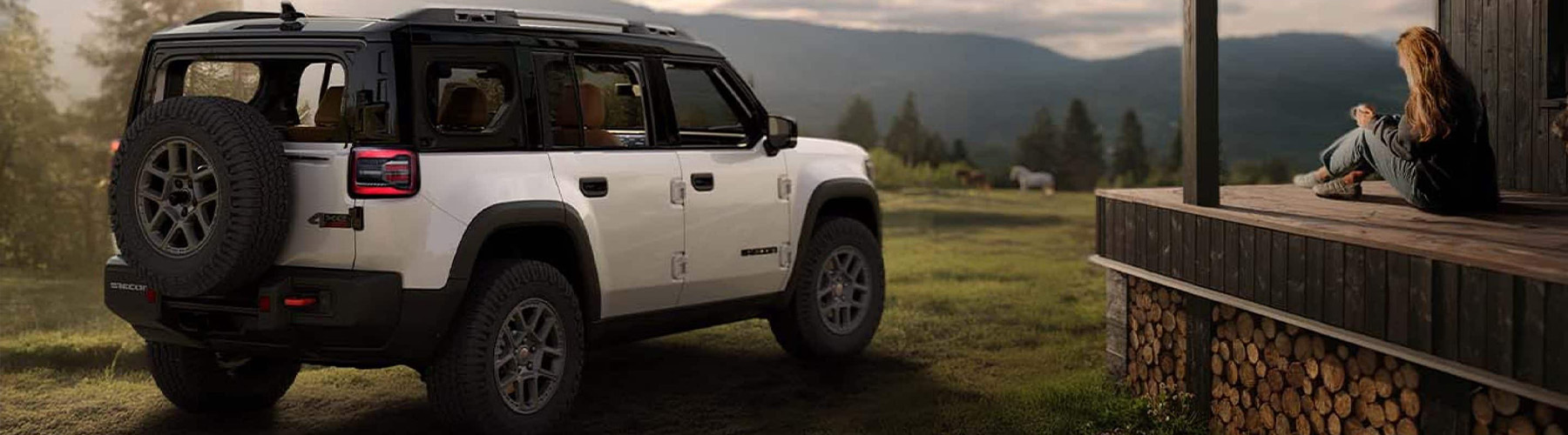 A white 2026 Jeep Recon with black roof, parked beside a large cabin with a woman and a dog on its porch and horses and mountains in the background.