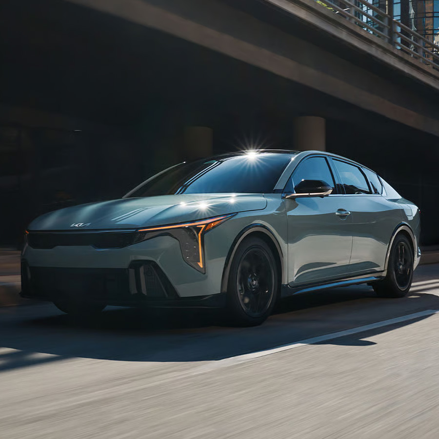 Driver-side front three-quarter view of the 2026 Kia K4 sedan GT-Line trim driving in an urban environment, featuring its unique front bumper with gloss black trim