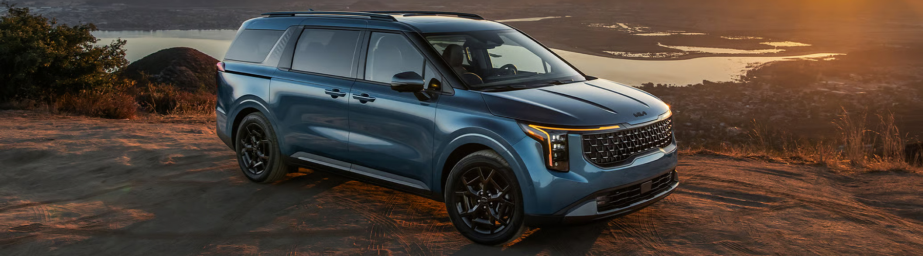 2026 Kia Carnival MPV in blue, SUV-inspired minivan, three-quarter side view, parked on a cliff overlooking the water at sunset