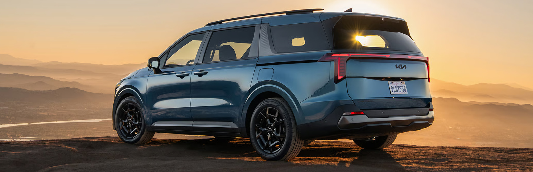 2026 Kia Carnival MPV in blue, SUV-inspired minivan, three-quarter back view highlighting the bold rear taillights, parked on a cliff at sunset