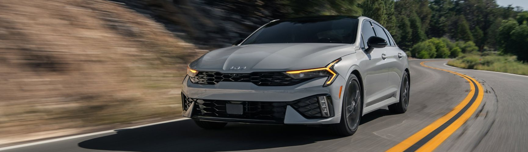 2026 Kia K5 Sedan in three-quarter front driver's side view, featuring the distinct headlights and design contours as it drives through sandy hillsides.