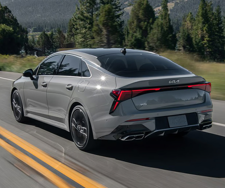 Rear three-quarter view of the 2026 Kia K5 Sedan driving confidently down a curving road, with tall trees and majestic mountainsides in the distance. The car shines in the sun, and the motion blur gives the photo a dynamic feeling.