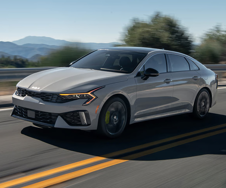  2026 Kia K5 Sedan, three-quarter driver's-side view, driving on a motion-blurred open roadway with scenic trees and mountains in the distance.