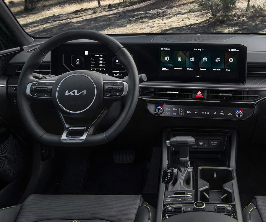 Interior dashboard, steering wheel, and console view in the 2026 Kia K5 Sedan, featuring the panoramic display, with trees visible through the windshield.