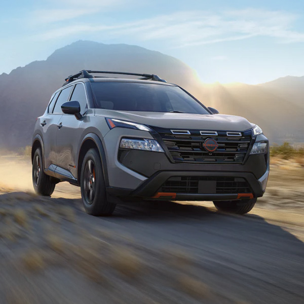 A dynamic shot of a gray 2026 Nissan Rogue SUV driving on a winding dirt road in a sunny, mountainous landscape, highlighting its rugged capability.