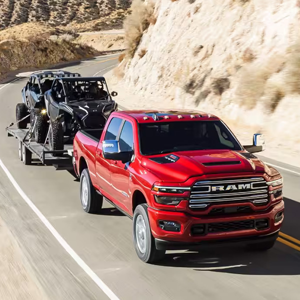 A red 2026 Ram 2500 Laramie 4x4 Crew Cab traveling on a highway in the mountains, while towing two ATVs.
